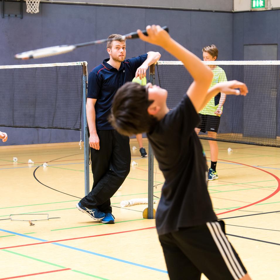 Gruppentraining im Badminton: Kinder üben mit Schlägern und Bällen unter Anleitung eines Trainers in einer Sporthalle.