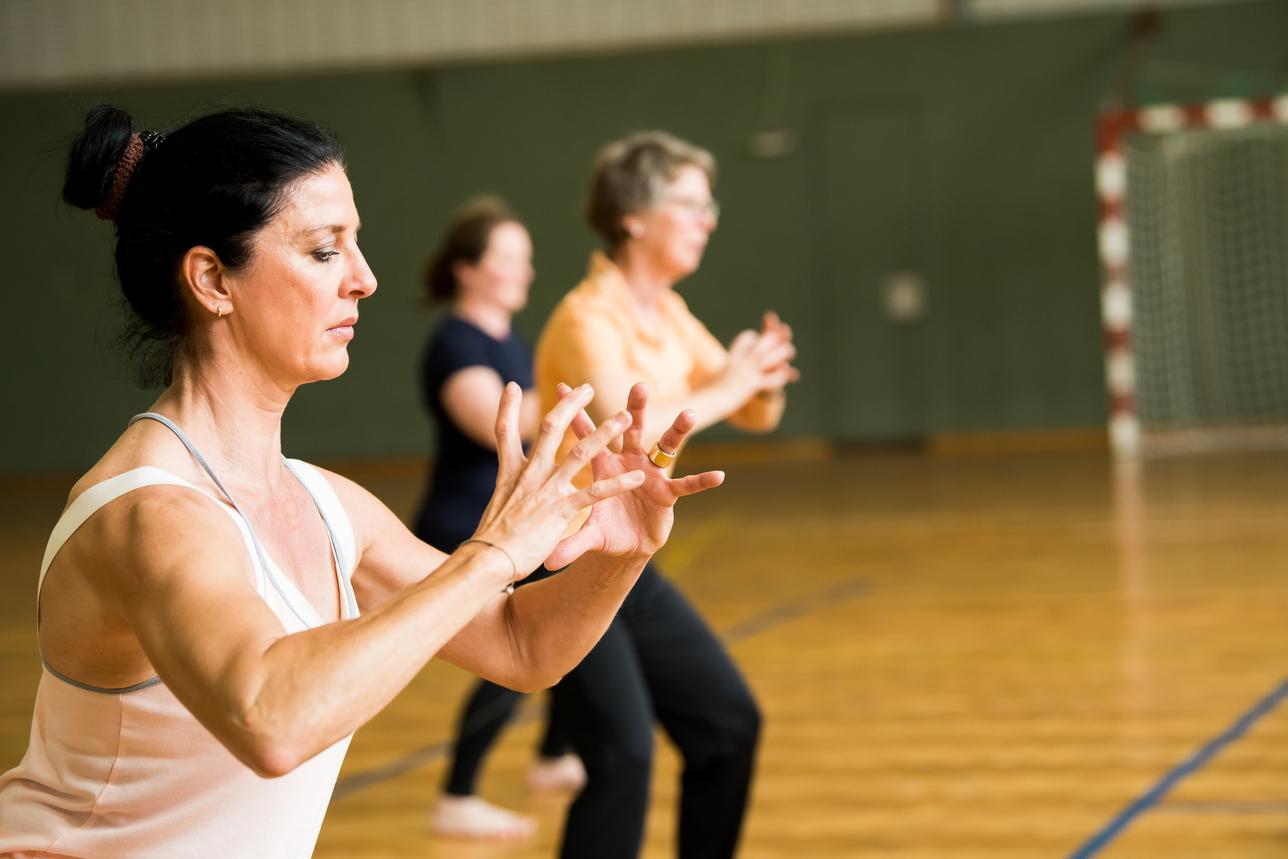 Drei Frauen führen eine Fitnessübung in einem Saal mit Holzfußboden und einer Sporttorwand im Hintergrund aus.