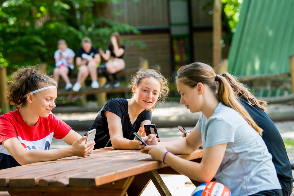 Vier Jugendliche sitzen an einem Holztisch im Freien und schauen auf ihre Smartphones.