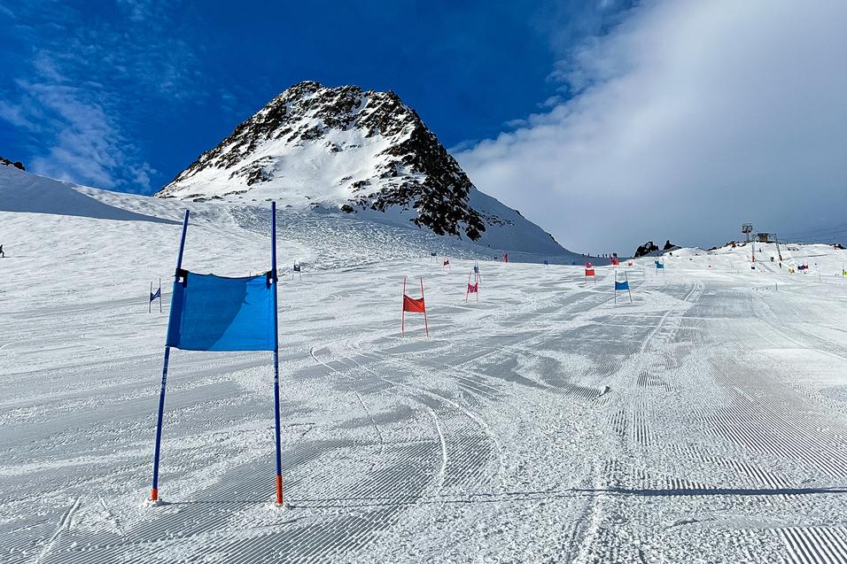 Skifahrer-Piste mit blauen und roten Toren, im Hintergrund ein schneebedeckter Berg unter blauem Himmel.