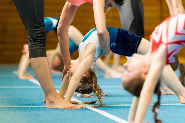 Trainer unterstützt ein Kind beim Rückwärtsspagat in einer Gymnastikhalle mit blauer Matte. Weitere Geräte im Hintergrund.